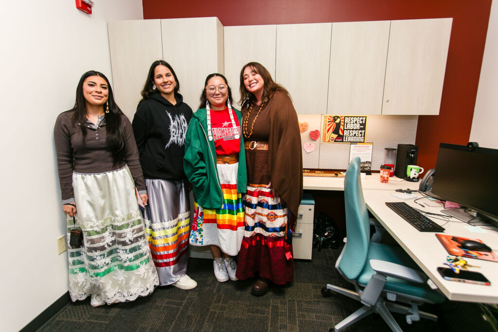 Four people standing inside an office area wearing ribbon skirts, with a desk and cabinets behind them.