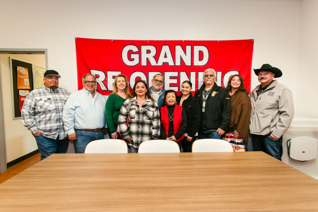 A group standing in front of a large red "Grand Re-Opening" banner inside a meeting room.