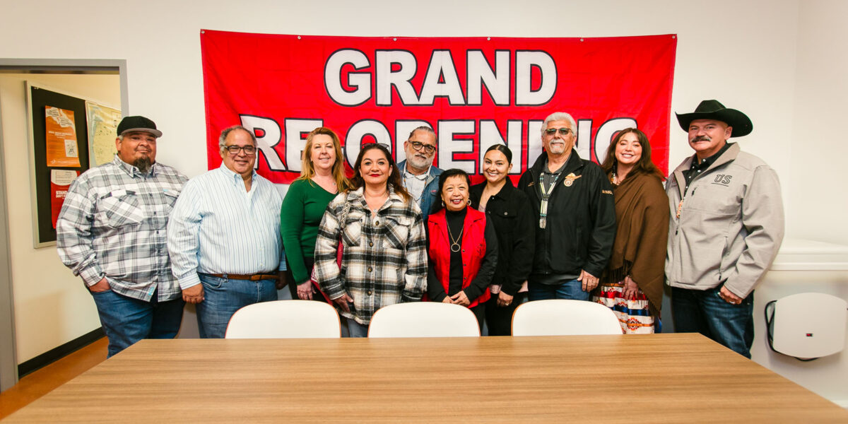 A group standing in front of a large red "Grand Re-Opening" banner inside a meeting room.