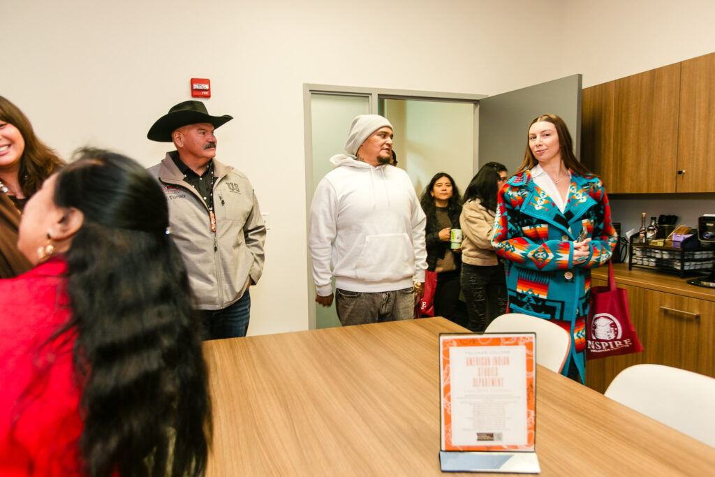 Several people standing in a room near a wooden table and break area, wearing a variety of coats and carrying red tote bags.