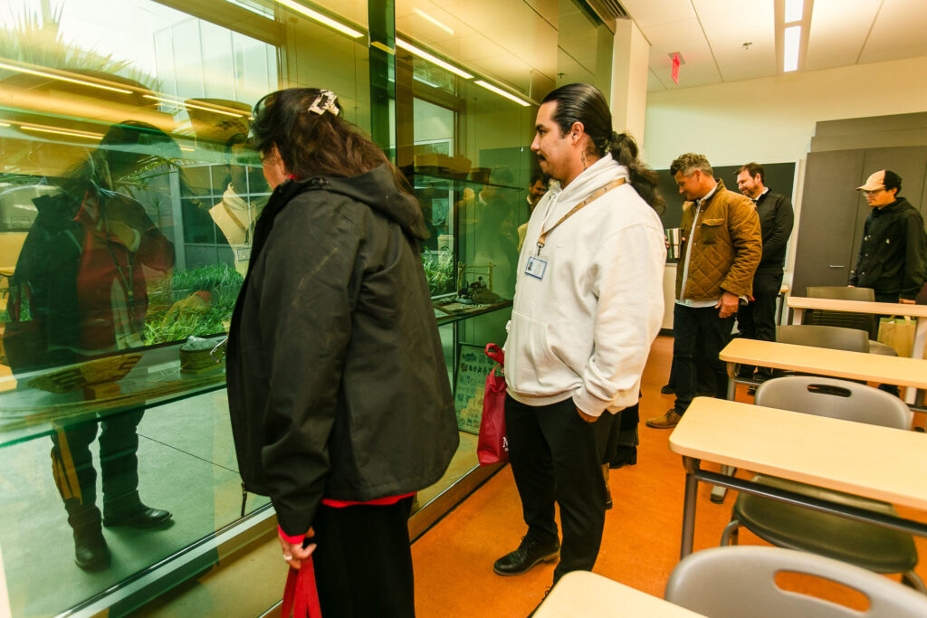 People inside a classroom standing near a large glass display containing cultural items, with desks visible in the foregrand.