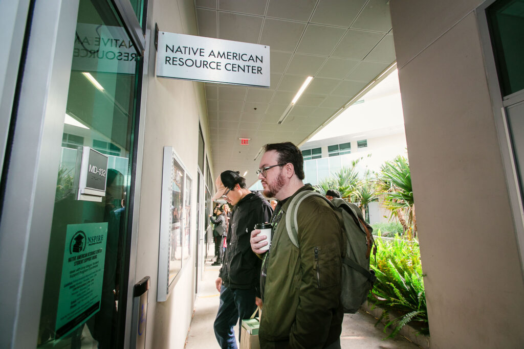 Two people standing outside near a concrete wall, one holding a cup, with natural light coming from the open walkway.