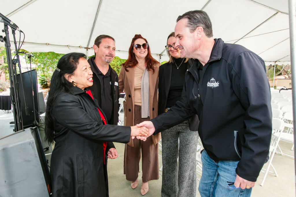 A group standing together under a white tent, featuring two people shaking hands with other attendees behind them.