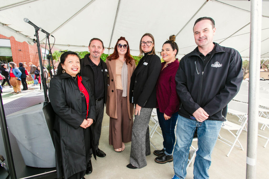 A group standing beneath an outdoor event tent arranged in a semicircle near the stage area.