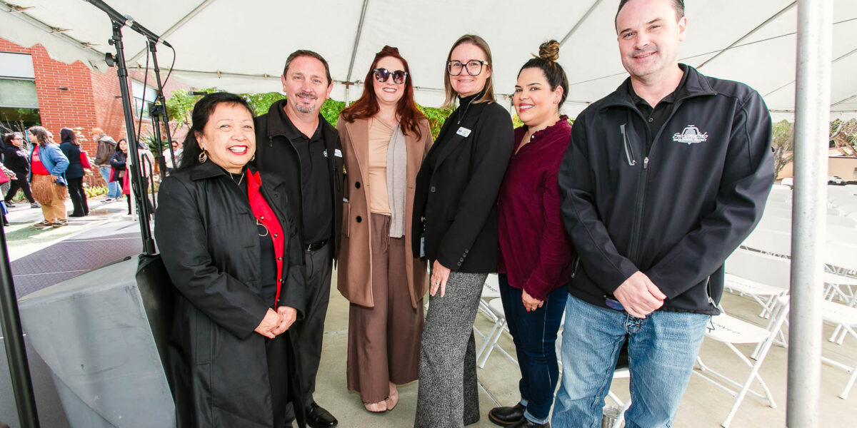 A group standing beneath an outdoor event tent arranged in a semicircle near the stage area.