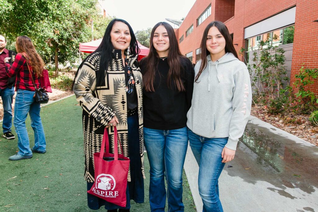Three people standing together outdoors near trees and a beige wall.