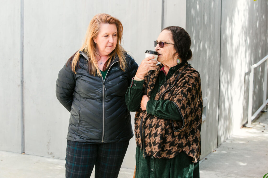 Two people standing on an outdoor walkway beside a concrete wall. One person holds a cup while chatting with the other person.