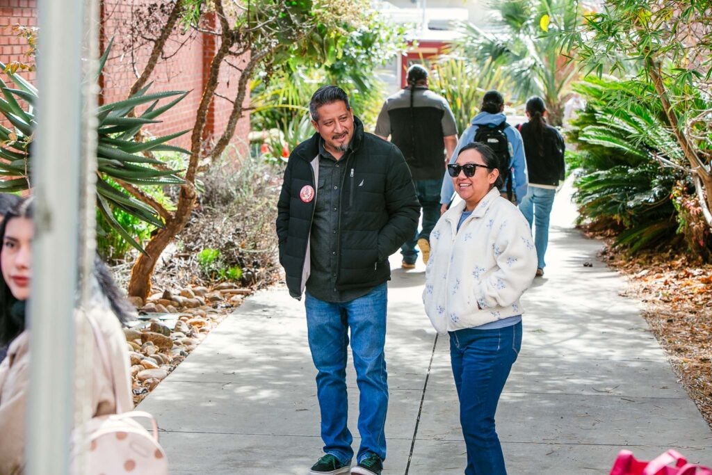 Multiple people walking along a concrete pathway lined with large leafy plants, small rocks, and trees.