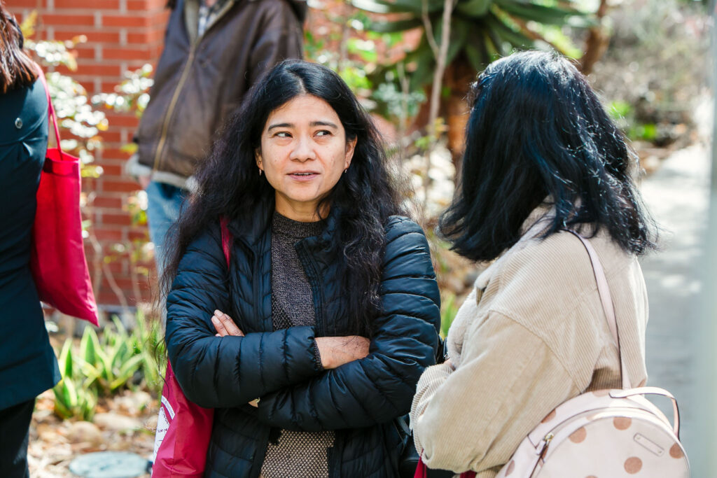 Two people standing on an outdoor walkway, with one person facing forward with folded arms, and another facing sideways with a backpack.