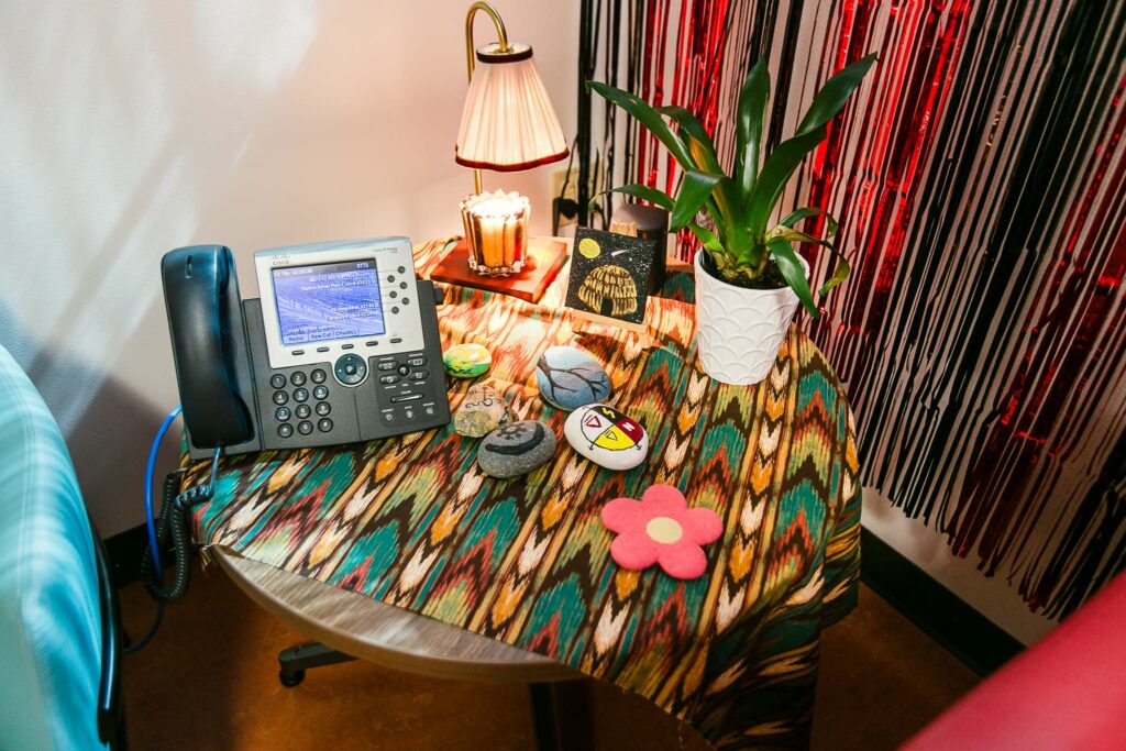 A small round table covered with patterned cloth holding a desk phone, painted stones, a lamp, a plant, and floral felted coasters.