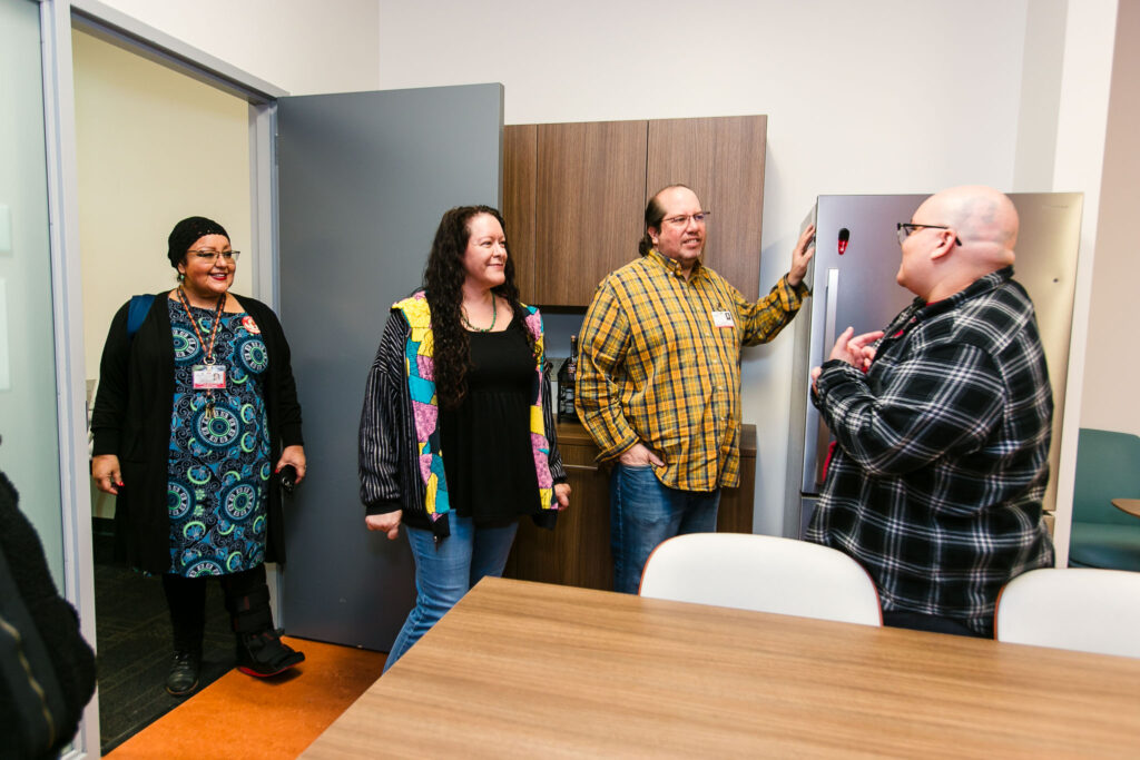 Several people standing in a kitchen space near cabinets and a refrigerator with a doorway open to another room.