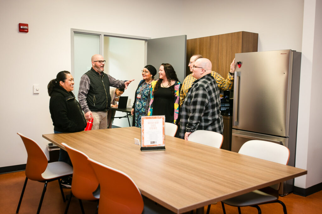 Several people standing inside a lounge space near a refrigerator and wooden cabinets, with a large table and chairs in the foreground.