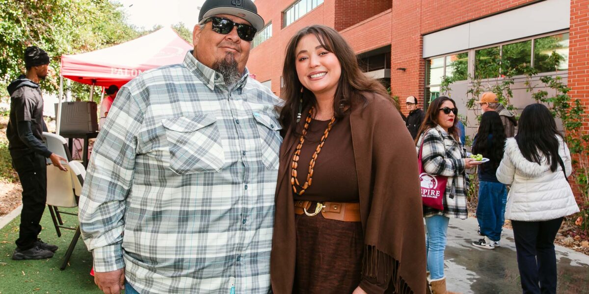 Two people standing side-by-side outdoors on artificial turf near a brick building.