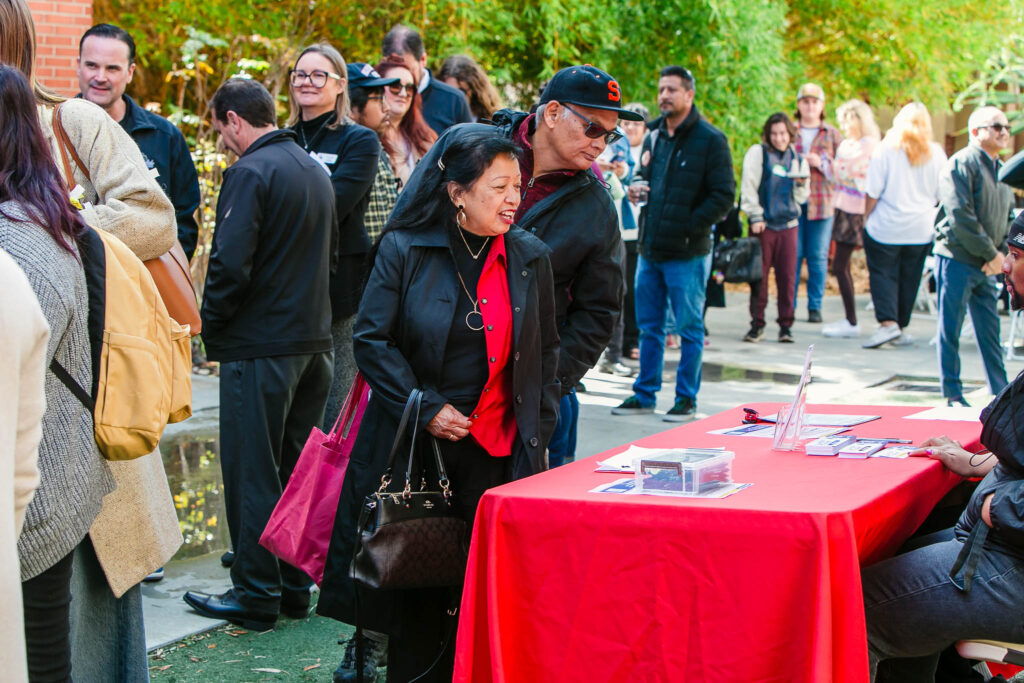 A line of people outdoors near a red-covered table holding informational materials and tote bags.
