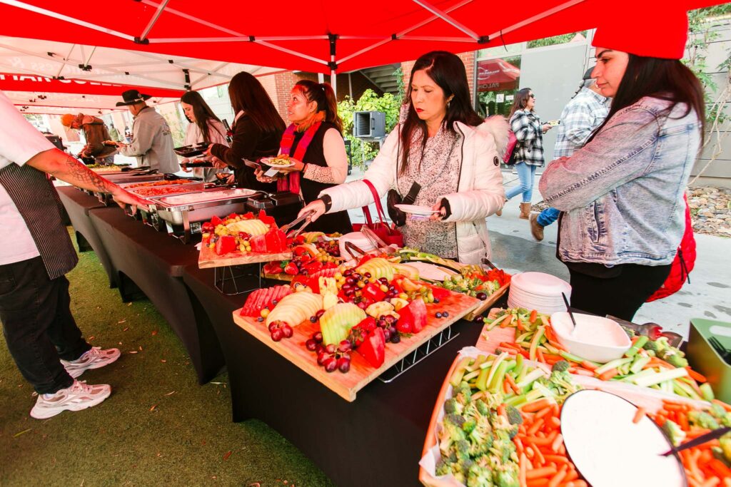 A large outdoor food table under a red canopy displaying fruit platters, vegetables, and serving utensils.
