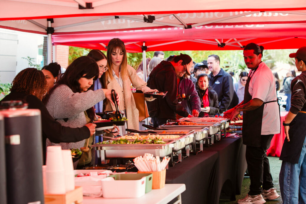 Several people standing under a red canopy selecting food from chafing dishes arranged on a long table.