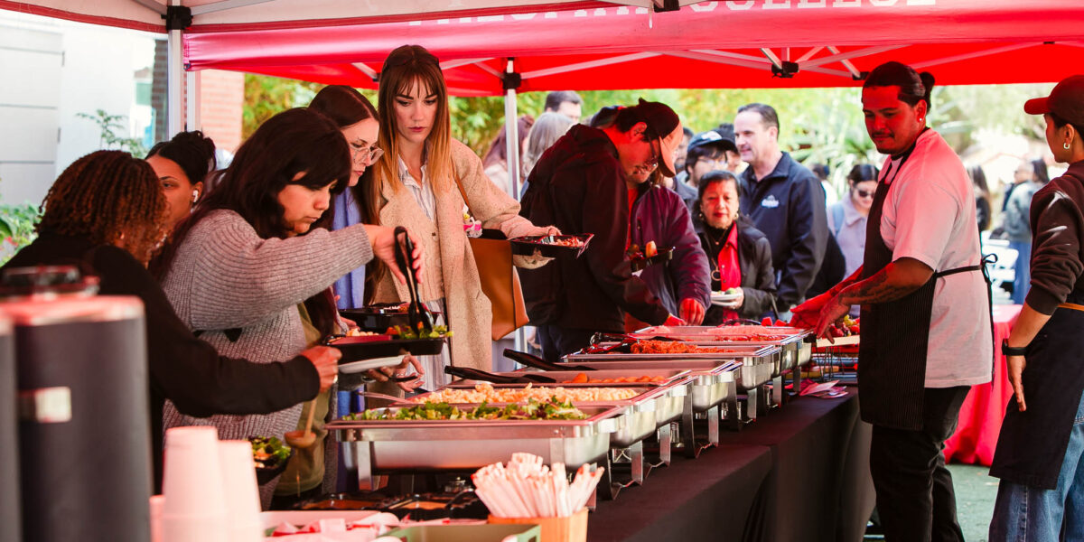 Several people standing under a red canopy selecting food from chafing dishes arranged on a long table.