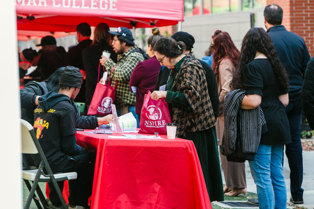 People standing in a line beside a red outdoor canopy. An information table covered with promotional materials.