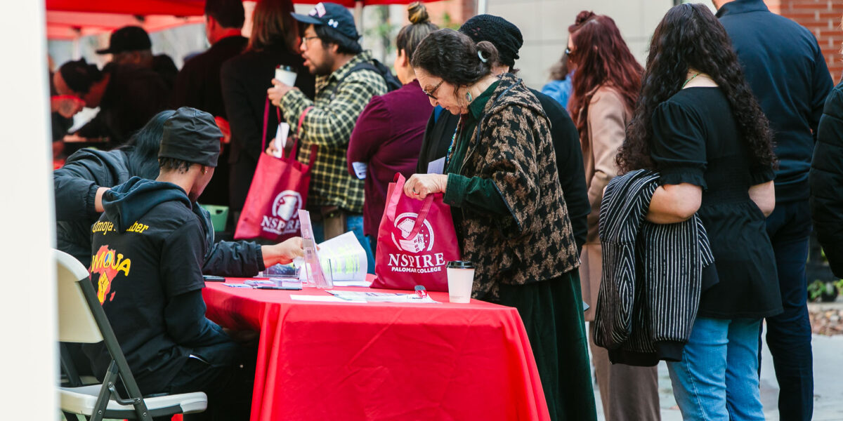 People standing in a line beside a red outdoor canopy. An information table covered with promotional materials.