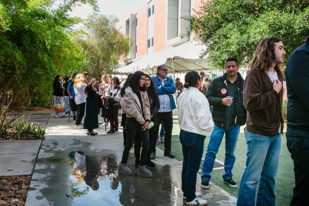 A long line of people standing outdoors on a walkway with tents and surrounding greenery.