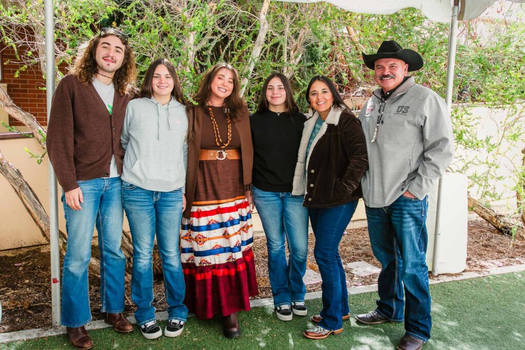 Six people standing under a canopy on artificial turf with plants and a wall behind them.