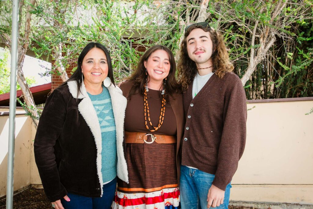 Three people standing closely together outdoors in front of a tall leafy shrubs and branches.