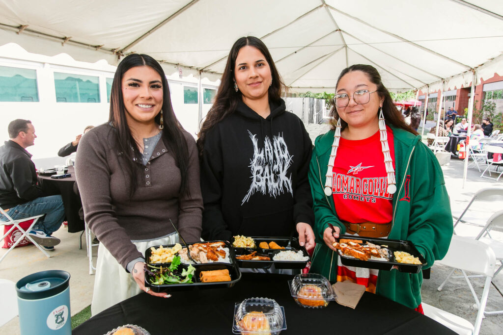Three people standing at a round table under a tent holding meal trays with assorted foods.