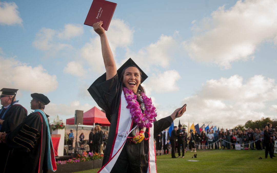 Female grade in cap and gown with a floral lei raising her diploma in the air