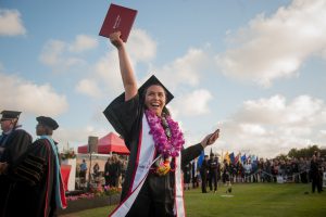 Female grade in cap and gown with a floral lei raising her diploma in the air