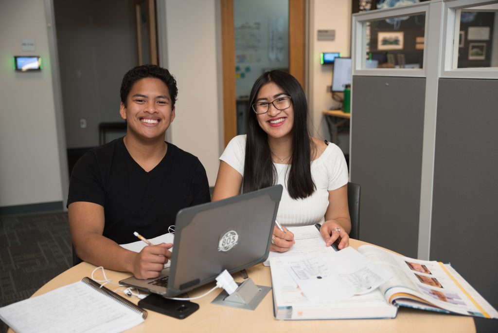 Two students sitting a table with a laptop, notes and textbook