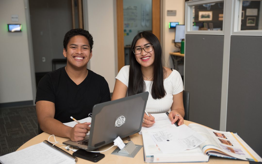 Two students sitting a table with a laptop, notes and textbook