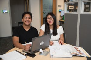 Two students sitting a table with a laptop, notes and textbook