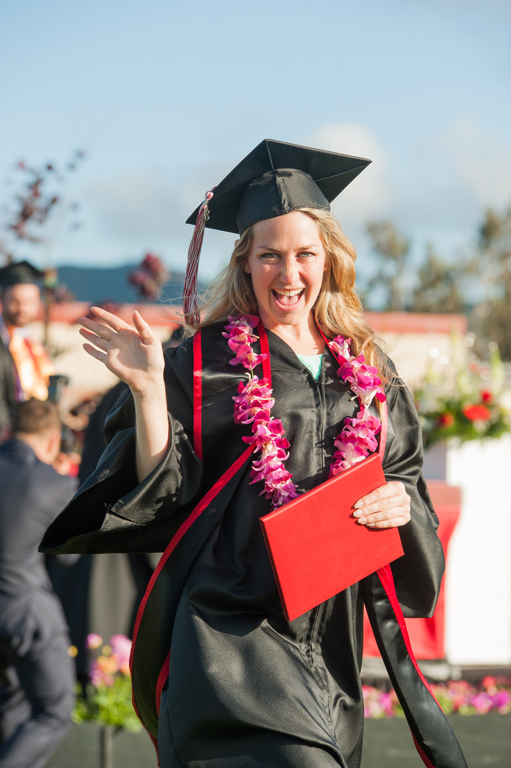 Female graduate in cap and gown and floral lei holding diploma and waving at commencement