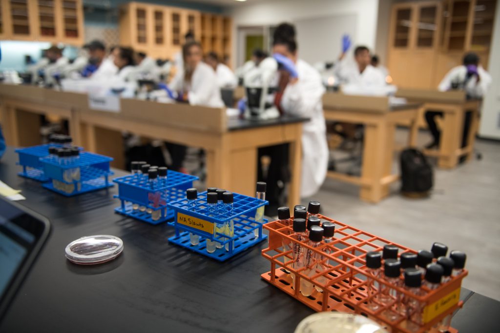 Biology class/lab with test tubes in foreground and students in lab coats at tables in the background