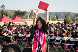 Excited Palomar grad wearing cap and gown and holding diploma up over her head