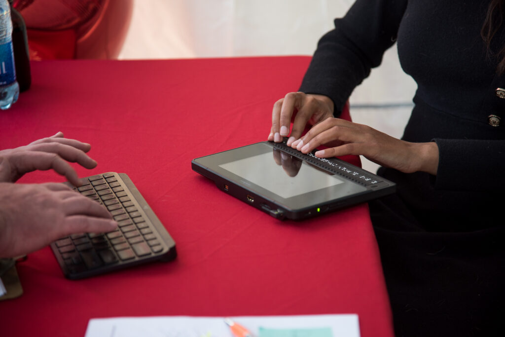 Two pairs of hands: one typing on a keyboard, one using a braille computer, on a red tablecloth