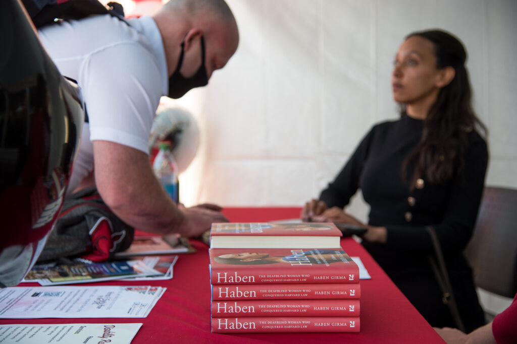 A stack of books entitled "Haben," while the author interacts with a student in the background