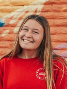 Dana is smiling in front of a orange, yellow, and blue brick wall wearing a red long sleeve shirt.