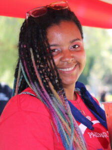 Chanti is in a red Palomar Promise shirt. She had rainbow dreadlocks and is smiling. 