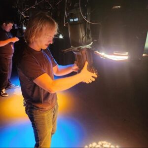 Two men are show wearing casual black clothing working on adjusting lighting instruments in a theatre.