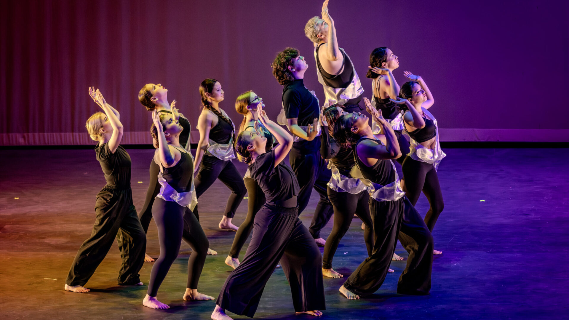 A photo from the production "Faculty Dance 2026". Onstage you see around 15 dancers moving to the right, all dressed in black, while the stage is filled with rainbow light.