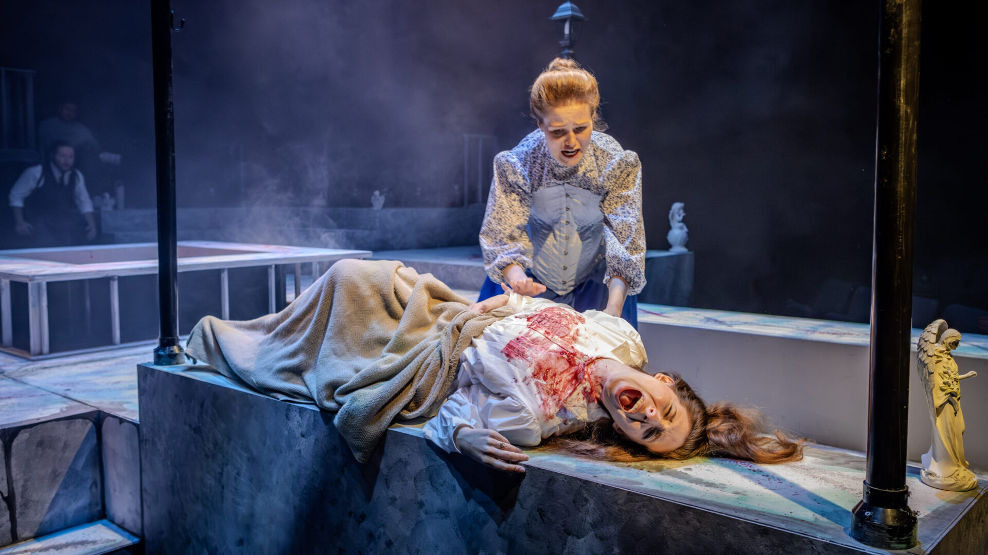 A photo from the production "Dracula, A Feminost Revenge Fantasy, Really". In a dark set stand two women, one lays down screaming covered in blood.