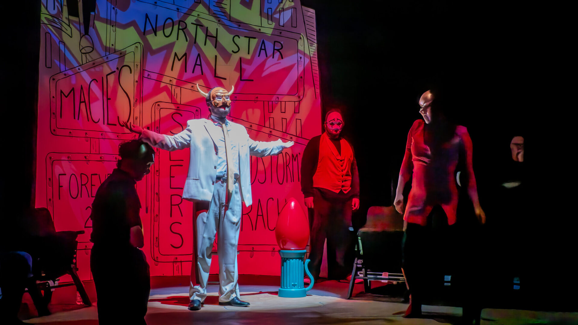 A photo from the production "Pastorela". A man in a white suit with an ambigous animal face mask stand, arms raised, preaching to group of people dress in formal black and red attire.