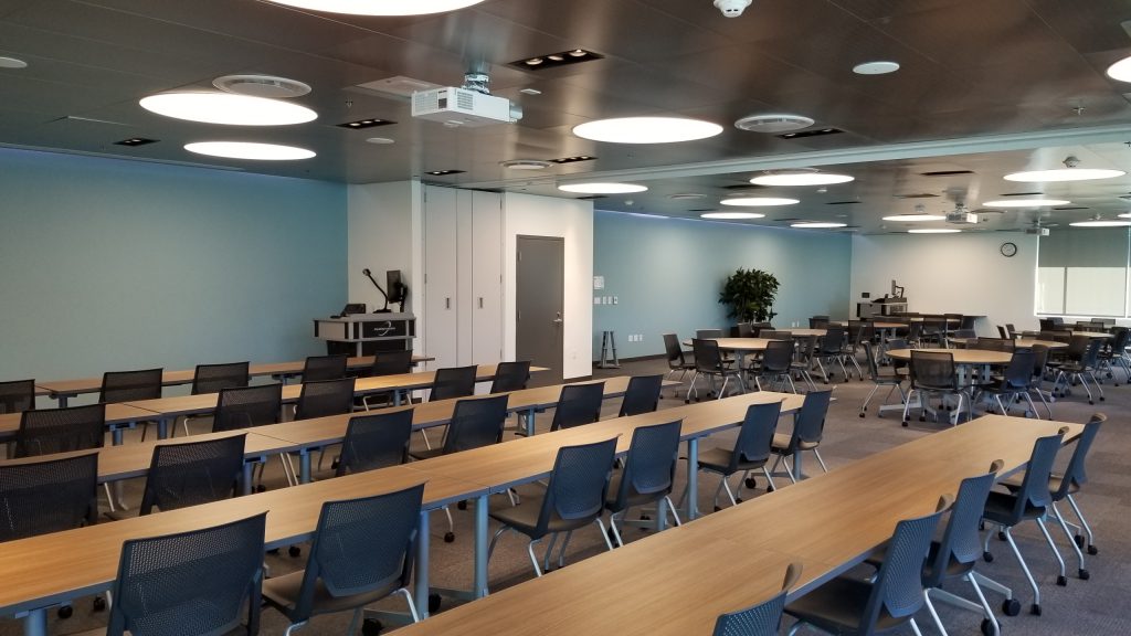 Rancho Bernardo Center Community Room. Room is empty but set up with tables and chairs in row on one side and chairs around round tables on the other side.