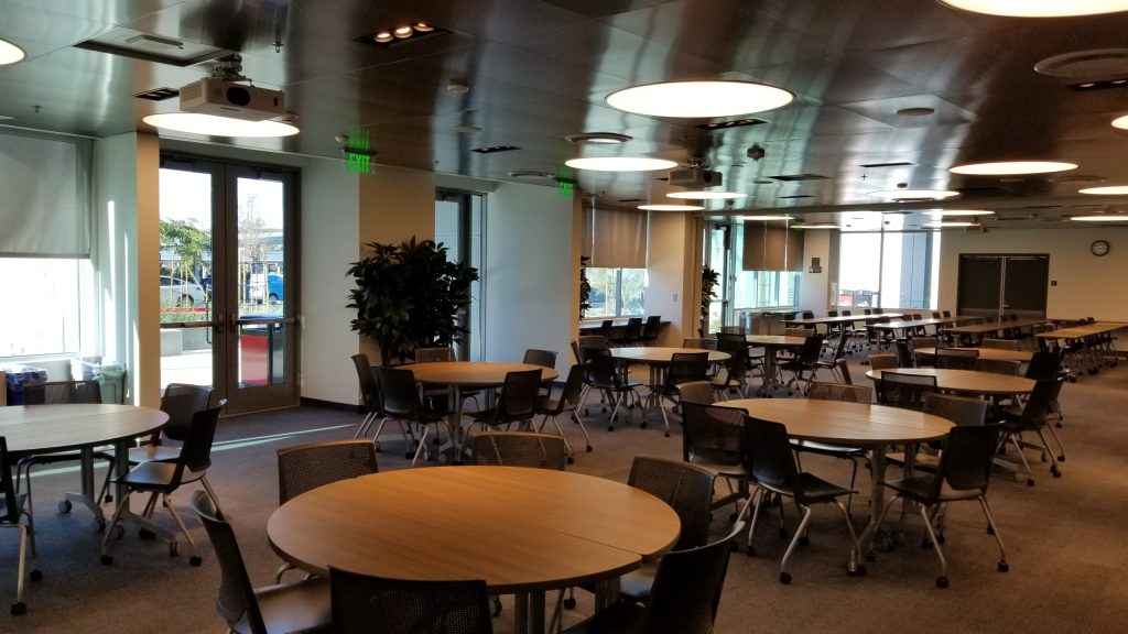 Rancho Bernardo Center Community Room. Room is empty but set up with tables and chairs in row on the far side and chairs around round tables on the near side.