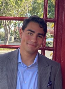 Elias is wearing a blue button up and grey suit jacket smiling in front of a red telephone booth.