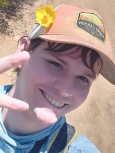 Lily is smiling at the camera, holding up a peace sign. She is wearing a blue shirt, orange hat, and yellow flower in their hair.