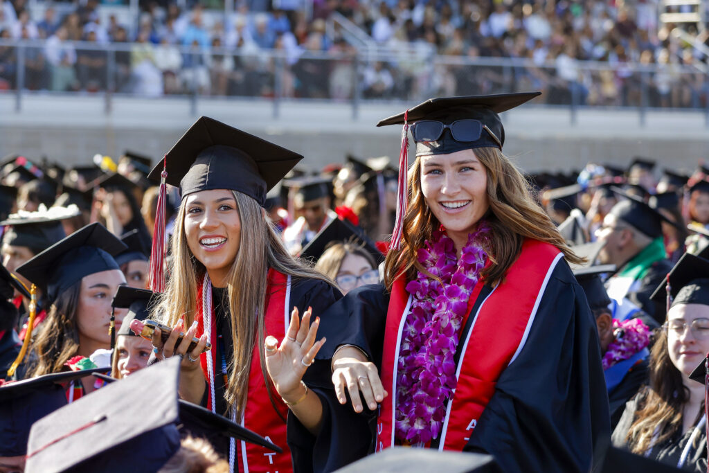 Two graduates in caps and gowns smiling and waving during a commencement ceremony, with fellow graduates seated in the background.