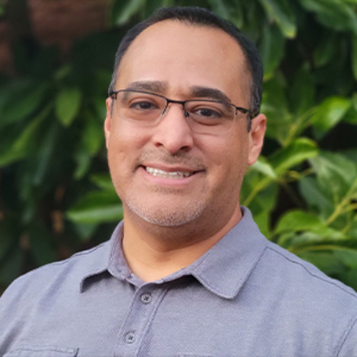 José Briceño is smiling and wearing black frame glasses and a blue gray collar polo. He is standing Infront of a leafy green backdrop.