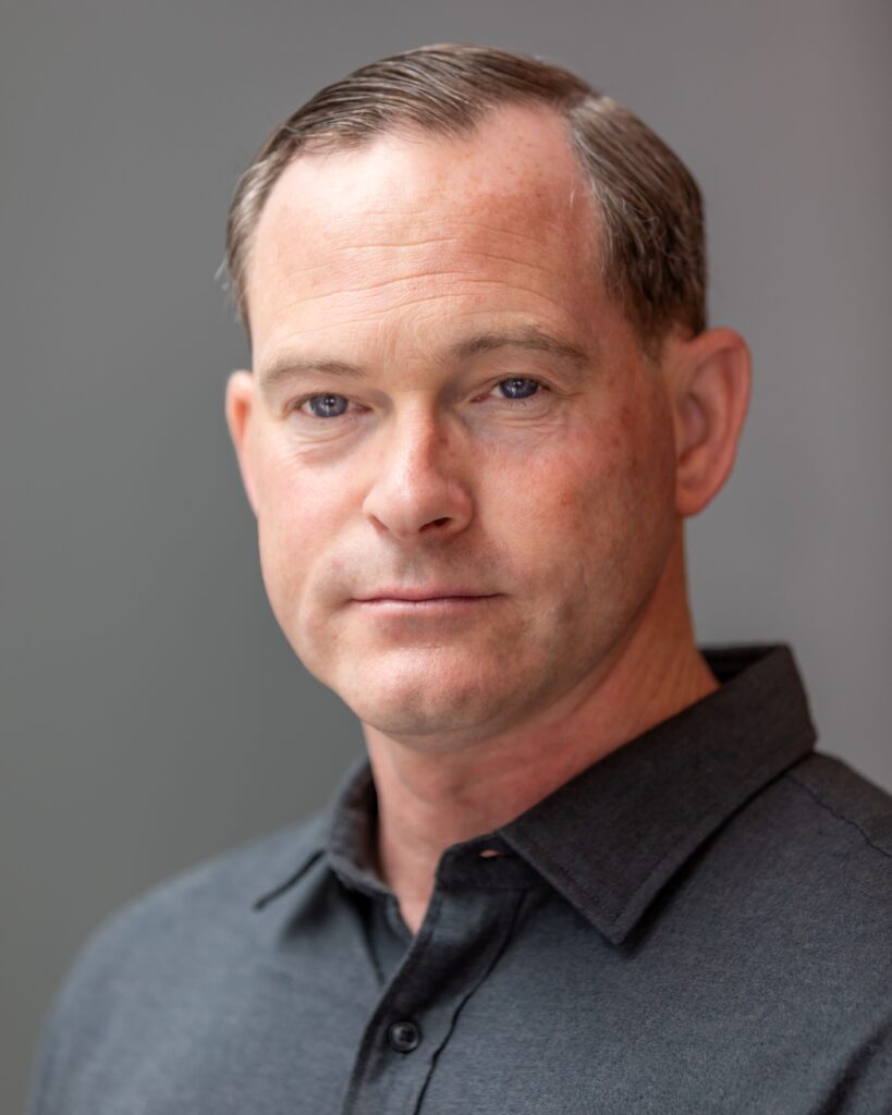 Daniel Balaszek is staring forward in front of a grey backdrop wearing a dark grey polo collared shirt.